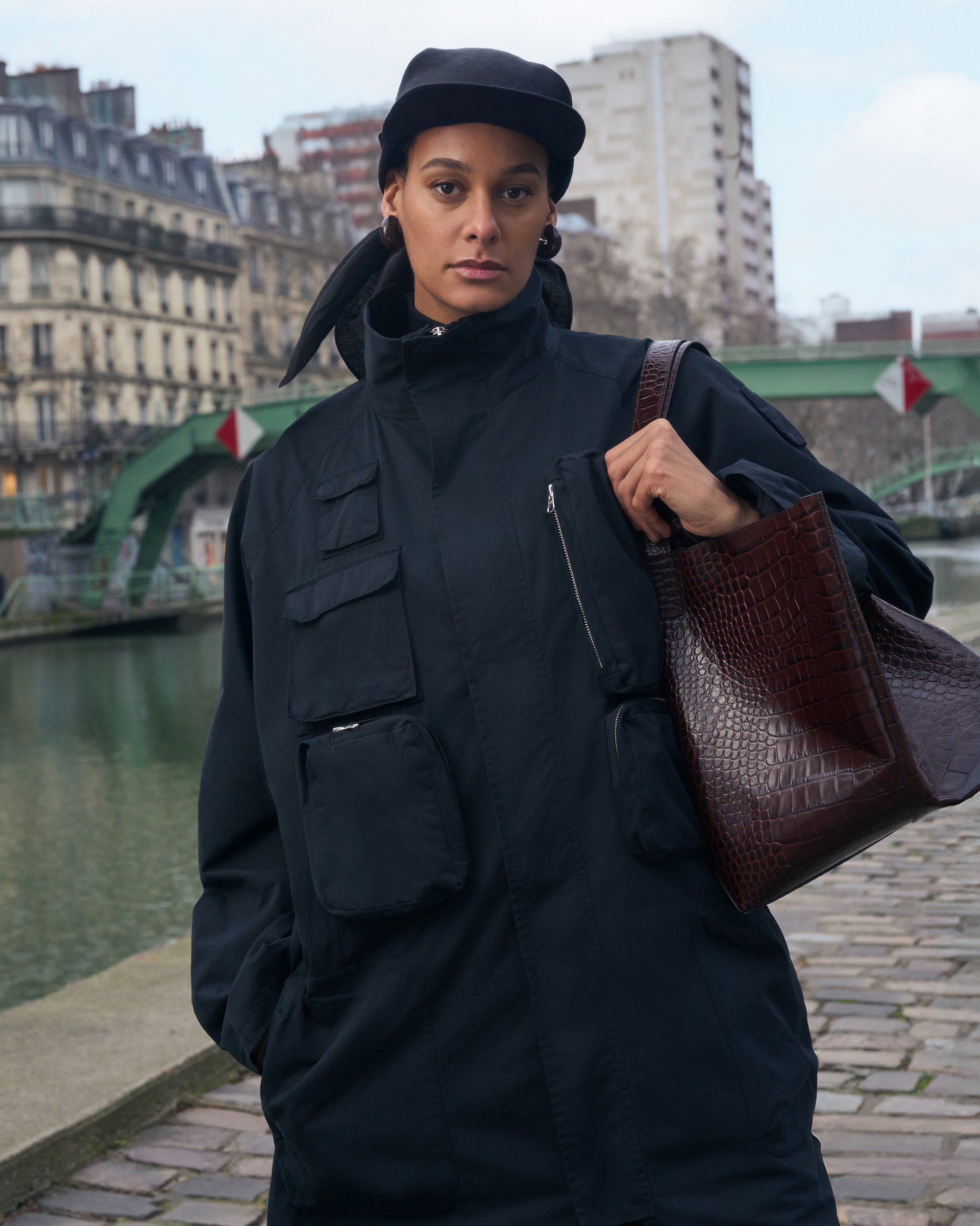 A woman in a black jacket and cap stands by a canal, holding a brown textured handbag, with a green bridge and city buildings in the background.