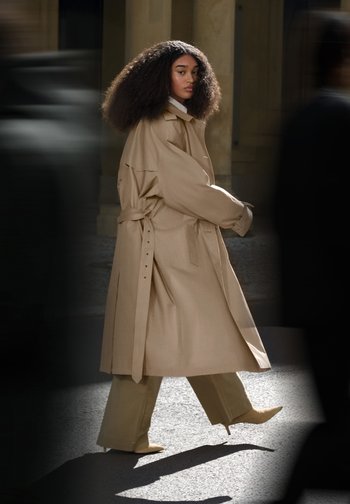 Beige trenchcoat met een oversized pasvorm en een ceintuur in de taille, gecombineerd met wijde crèmekleurige broeken en puntige beige hakken. Zacht, gestructureerd materiaal.