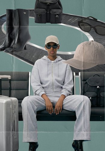 Henkilö, jolla on beige lippalakki, mustat aurinkolasit, harmaa verkkapuku, istuu lentokentän penkillä hopeanvärisen matkalaukun, mustien saappaiden ja reppujen ympäröimänä.