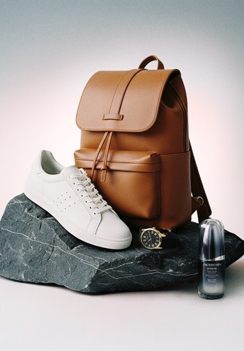 Brązowy skórzany plecak, biały sneaker, czarno-złoty zegarek na nadgarstek i butelka serum Shiseido Men Ultimune na ciemnej kamiennej płycie.