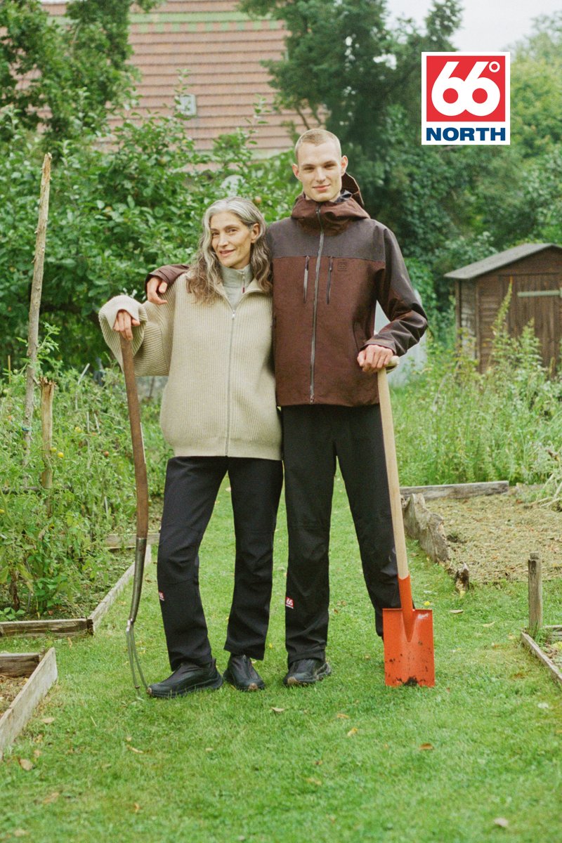 Due persone sono in piedi in un giardino, indossando un maglione spesso beige e una giacca marrone con zip, insieme a pantaloni neri e stivali. Tengono una falce e una pala.