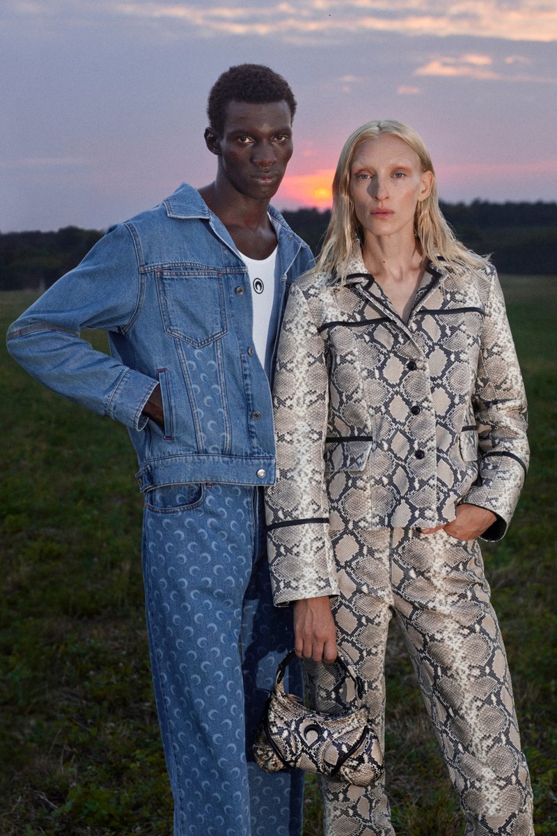Two models stand in a field near a sunset. One wears a denim jacket and patterned pants, while the other sports a snake-print suit and handbag.