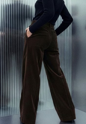 Bruine corduroy broek met wijde pijpen, zijzakken en verticale ribbelstructuur, gedragen met een nauwsluitende zwarte top tegen een gestructureerde achtergrond.