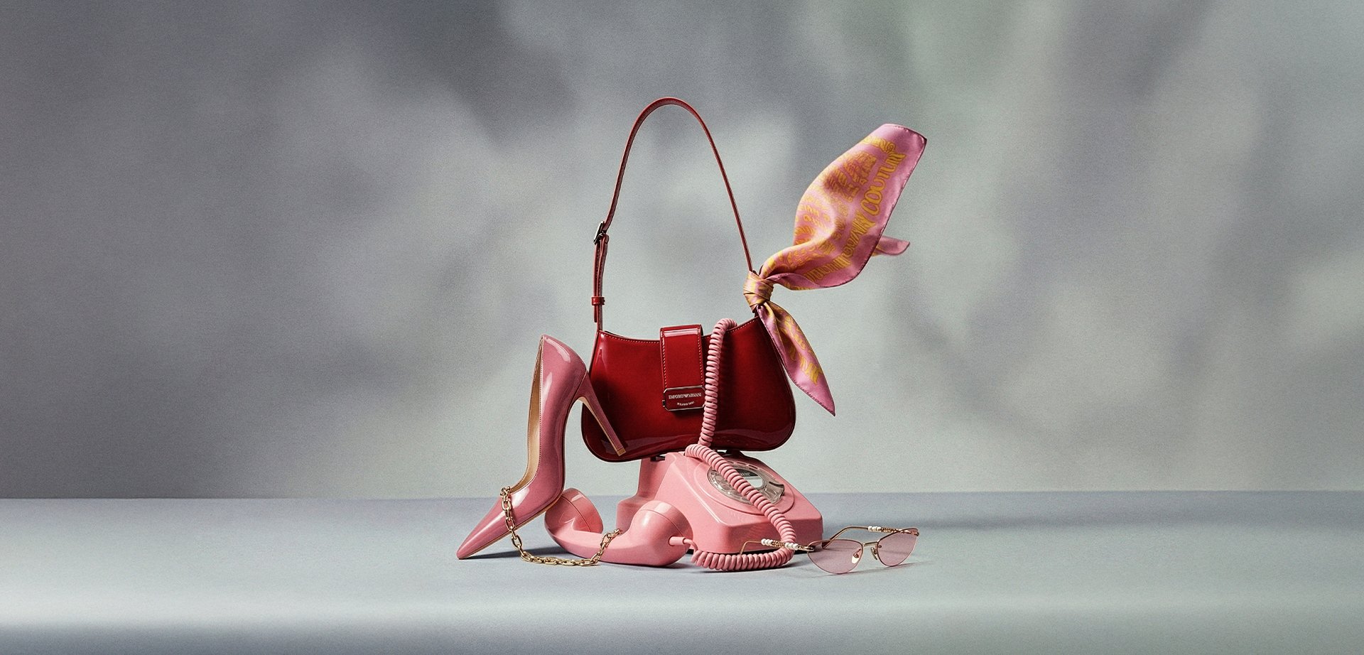 Pinkfarbener Wählscheibentelefon mit gewendeltem Kabel, rote Handtasche mit an den Griff gebundenem pinkfarbenem Schal, pinkfarbene High-Heels mit Kette und rosa getönte Sonnenbrille.