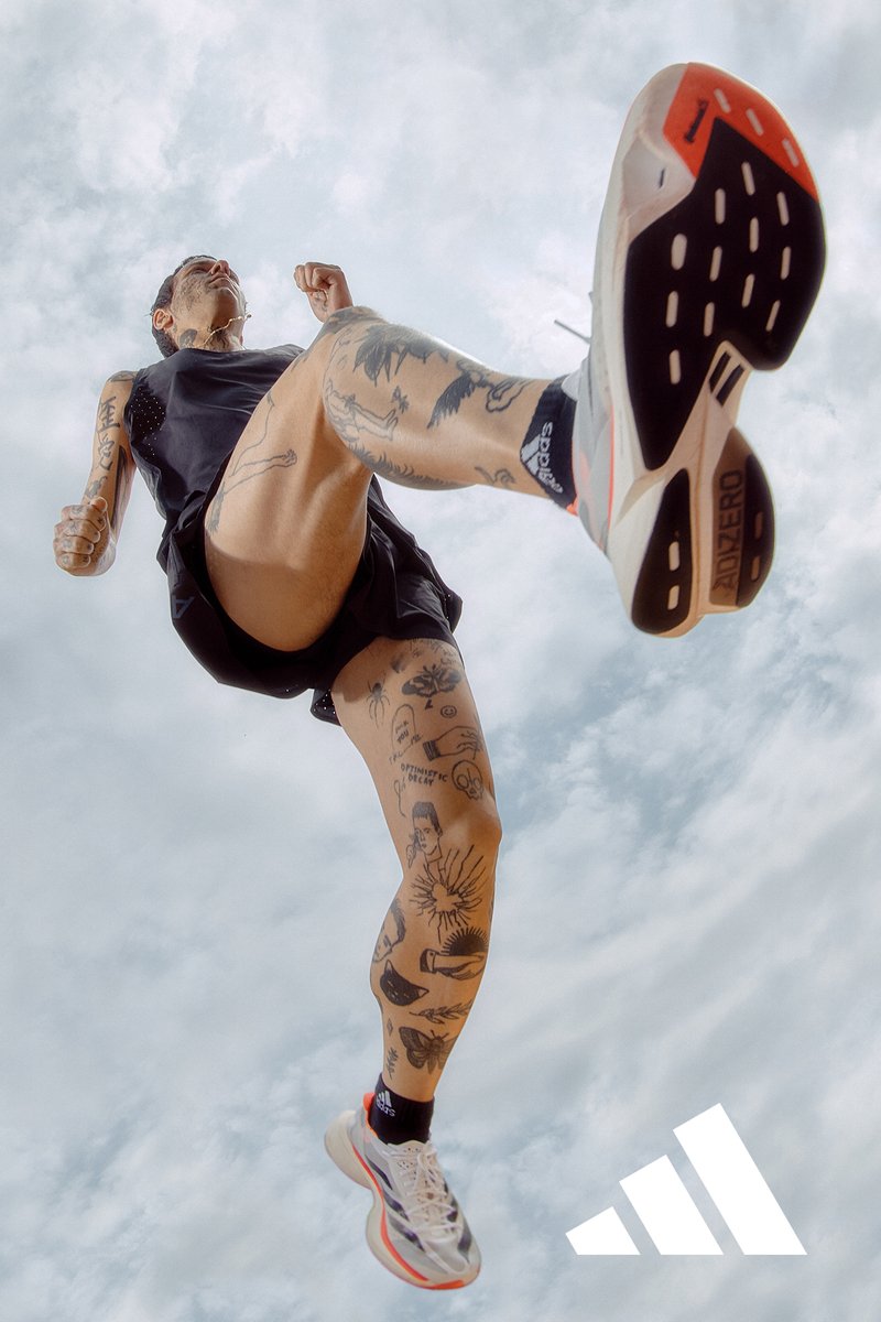 Athlete in motion, running with tattoos visible on legs. Wears black shorts and brightly colored shoes against a cloudy sky backdrop.