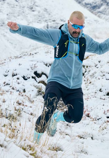 Αέρινο, μπλε αδιάβροχο μπουφάν πάνω από ένα μαύρο γιλέκο, μαύρη βερμούδα καιτιρκουάζ παπούτσια. Χιονισμένο έδαφος και ξηρά χόρτα στο υπόβαθρο.