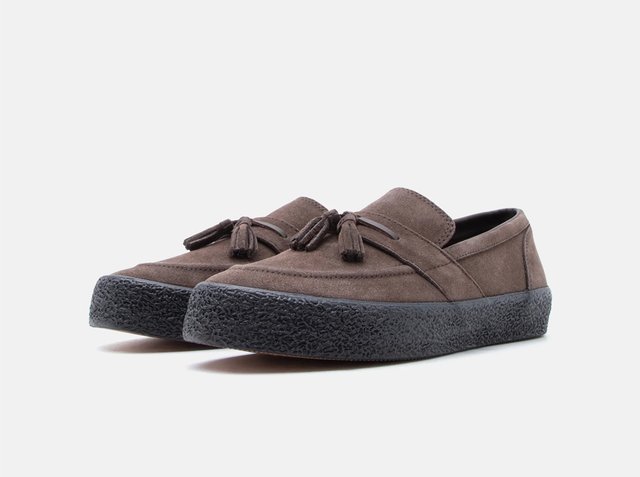Brown suede slip-on shoes with black textured rubber soles and decorative tassels on the top, shown from a low side angle.