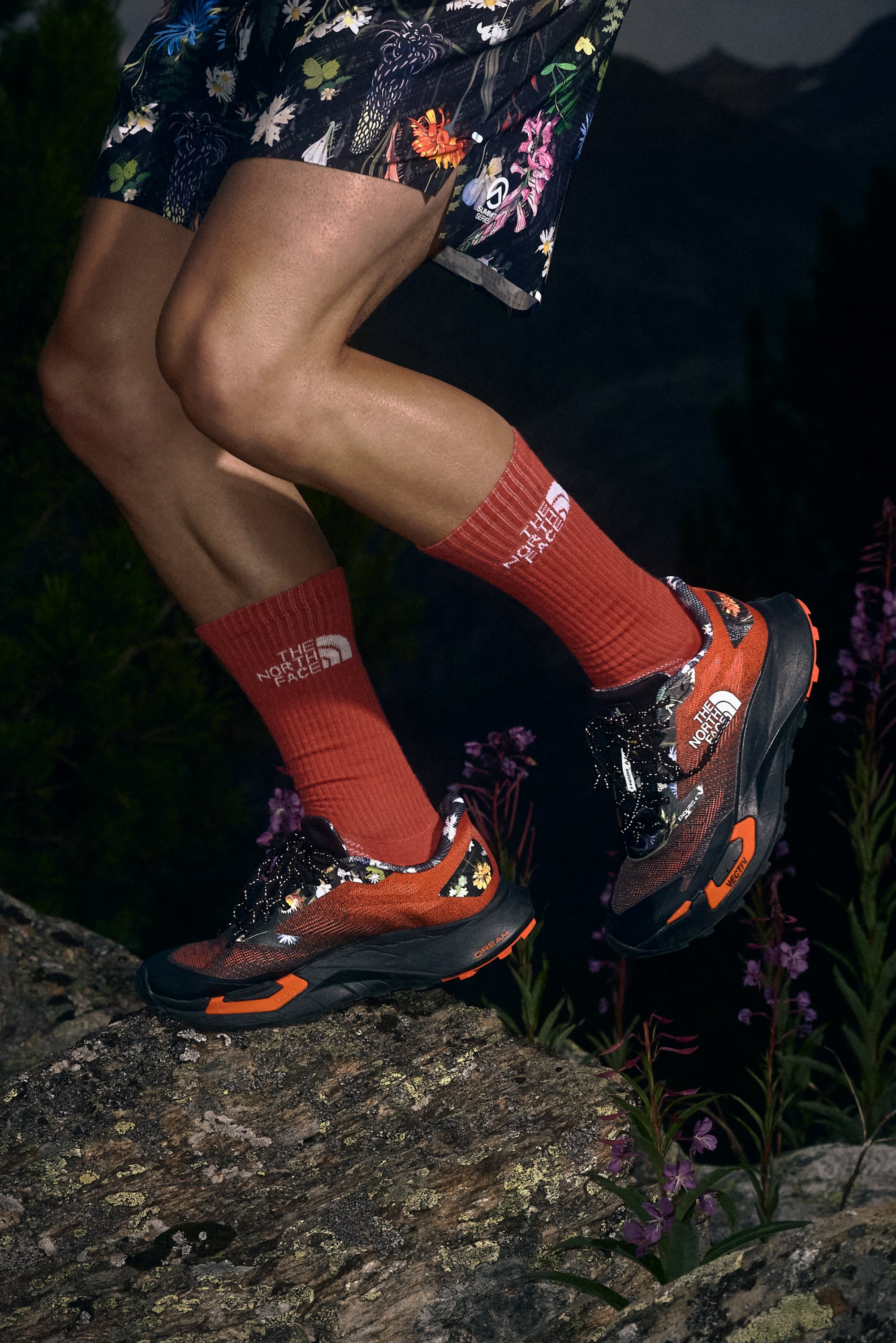 Zapatillas de trail running rojas y negras con acentos florales, empeine de malla texturizada, suela de goma y cordones negros. Llevadas con calcetas rojas de marca.