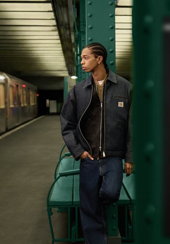 Mann mit geflochtenem Haar, der eine dunkle Jacke und Jeans trägt, lehnt an einer grünen Metallstütze auf einem leeren U-Bahnsteig, im Hintergrund ein Zug.