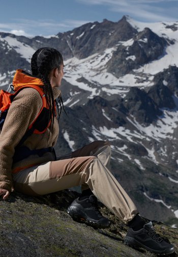 A woman with braided hair sits on a rocky outcrop, overlooking snow-capped mountains, dressed in outdoor attire with an orange backpack.