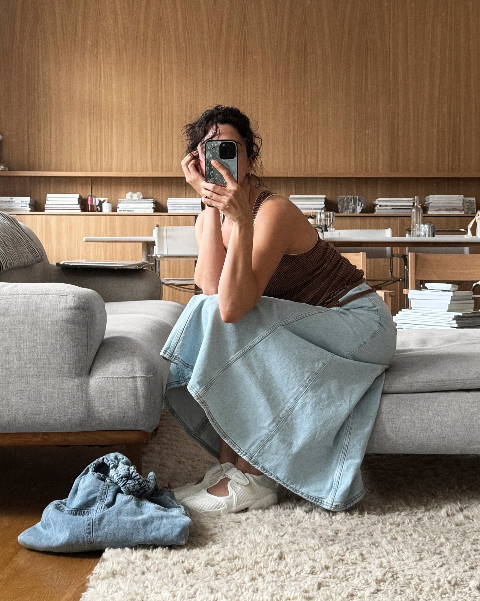 Una mujer está sentada en un sofá, sosteniendo un teléfono y sonriendo a la cámara. Lleva una blusa marrón y una falda de mezclilla azul, con un par de zapatillas cerca.