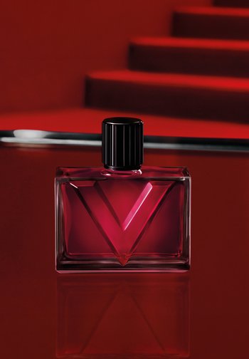 Flacon de parfum en verre rouge avec bouchon noir sur surface rouge brillante, grand motif rouge "V" à l'avant, escaliers rouges flous en arrière-plan.