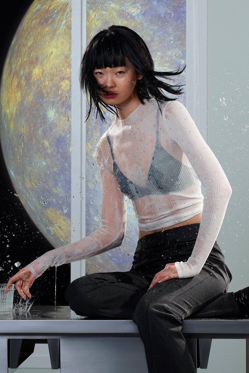 Una joven con el cabello mojado, vistiendo una blusa transparente y pantalones oscuros, se sienta en una mesa salpicando agua, frente a un fondo cósmico.