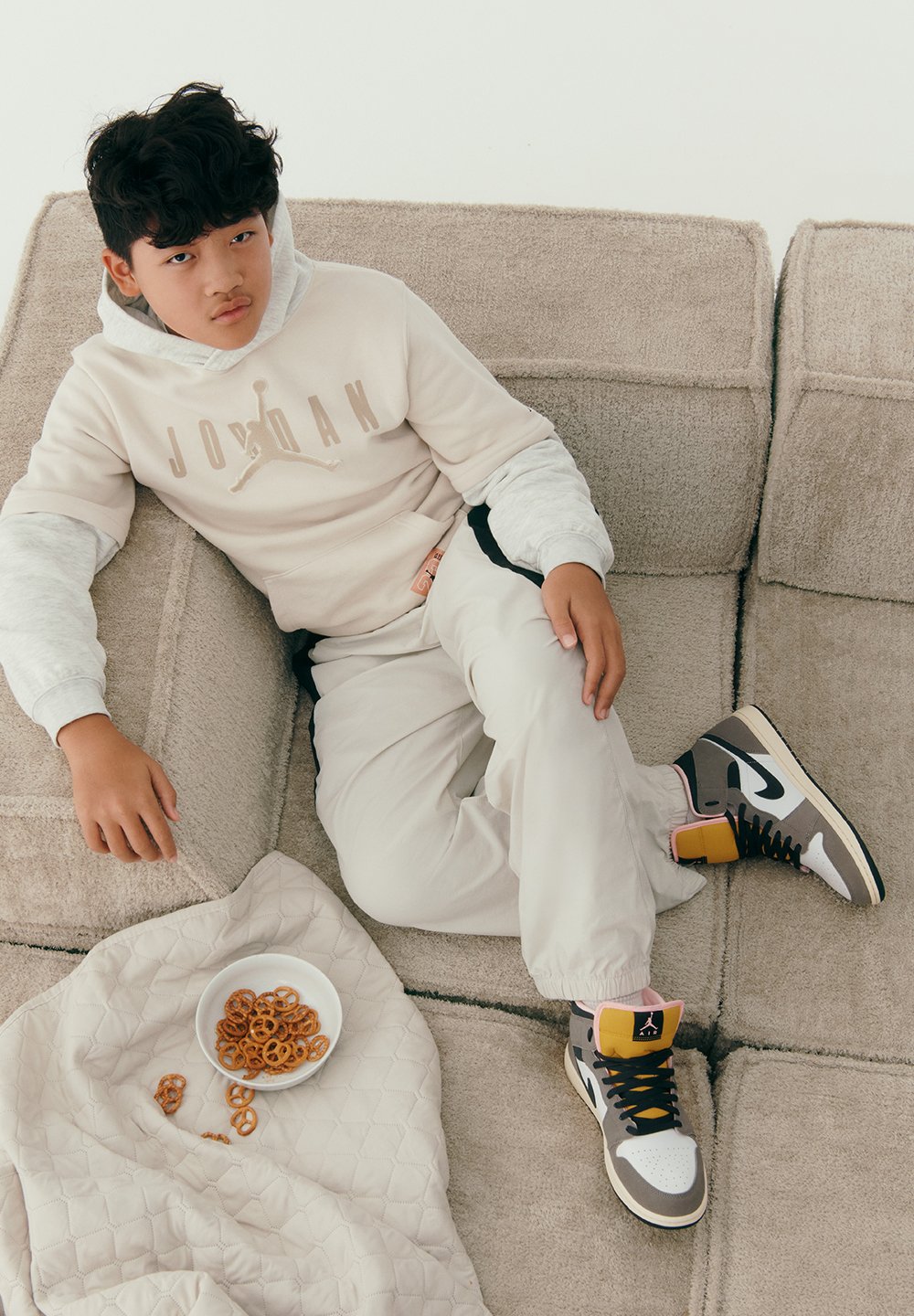 Sudadera Jordan en beige con logo, mangas gris claro; pantalones crema; zapatillas altas multicolores con acentos en negro, beige y amarillo. Plato blanco de pretzels.