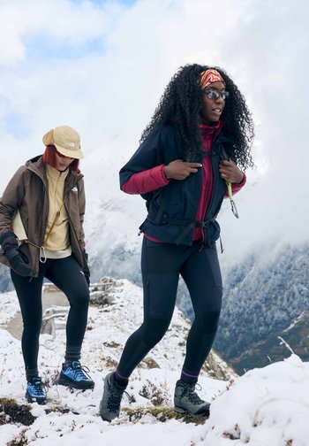 Δύο πεζοπόροι ανεβαίνουν σε ένα χιονισμένο μονοπάτι. Ο ένας φοράει ένα καφέ μπουφάν και μια μπεζ καπέλο; ο άλλος έχει ένα σκούρο μπουφάν με μπορντό επένδυση και μια σακίδια.