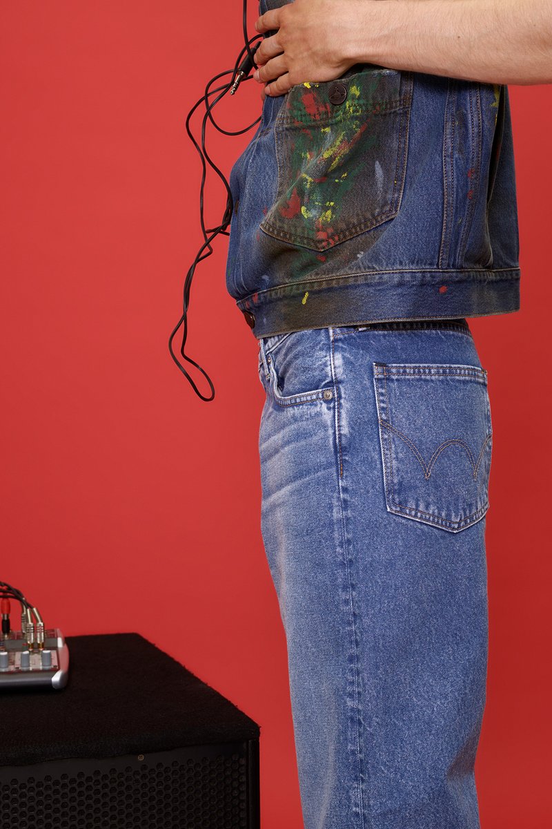 Persona con chaqueta de mezclilla salpicada de pintura y jeans azules, sosteniendo un cable de audio cerca de un mezclador de sonido sobre un altavoz negro contra un fondo rojo.