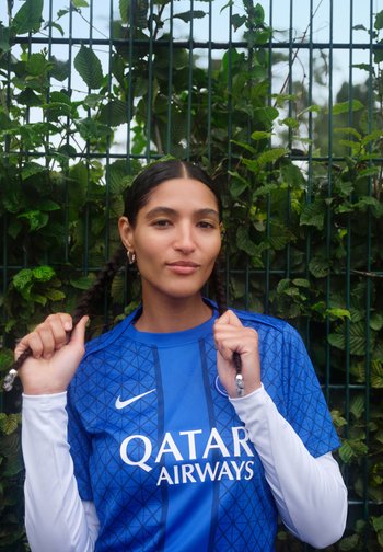 Noor naine sinises Qatar Airways jalgpallijerseys, kes hoiab punutud patsikest, seismas rohelise lehtedega aia ees õues.