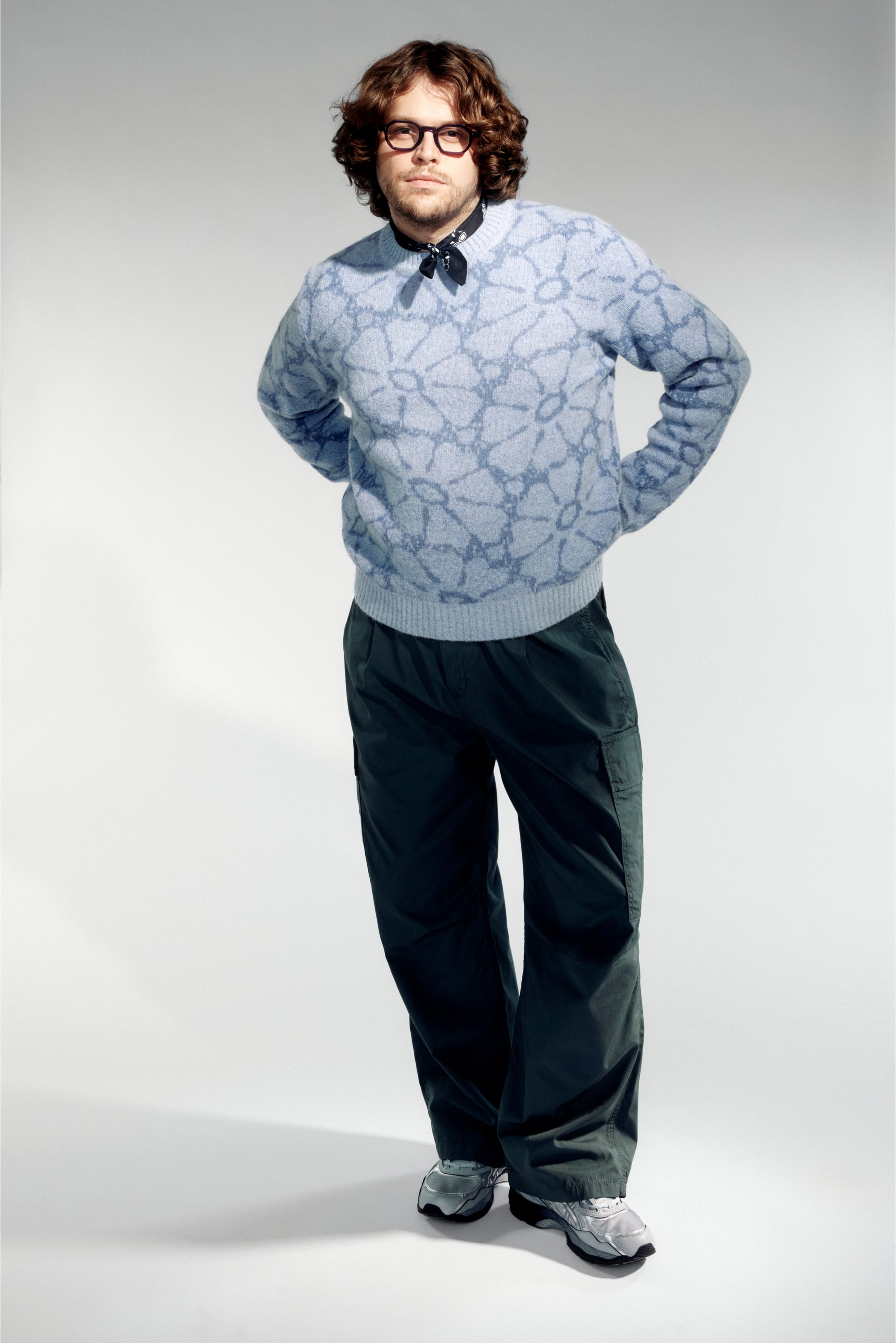 Hombre con cabello rizado que lleva gafas, un suéter de patrón azul claro, pantalones negros y zapatillas, de pie con las manos en las caderas contra un fondo gris.