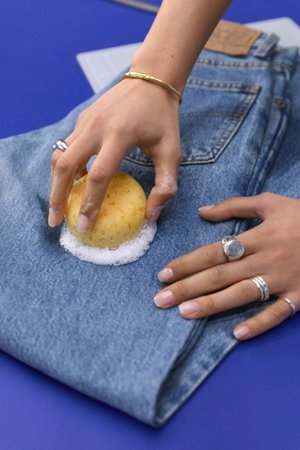 A hand scrubs a soapy sponge on a folded pair of blue denim jeans, with a second hand displaying multiple rings nearby.