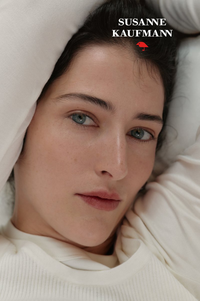 Nærbillede af en kvinde med blå øjne og naturlig makeup, iført en hvid top, der hviler sit hoved på hvidt stof.