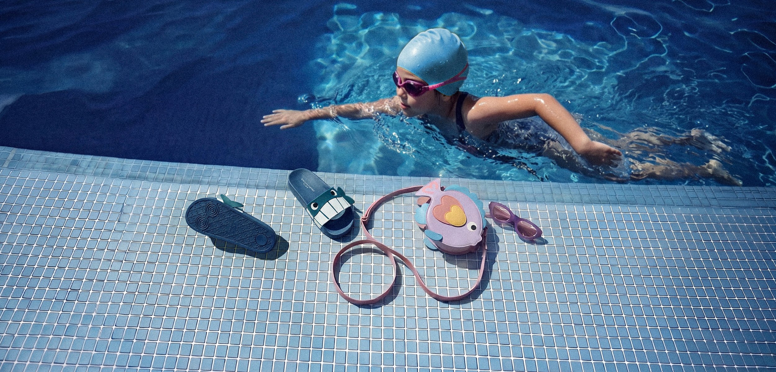 Kind mit Badekappe und Schwimmbrille schwimmt in einem Pool nahe dem Rand mit Sandalen, taschenförmiger Tasche und Sonnenbrille auf dem gefliesten Poolbereich.