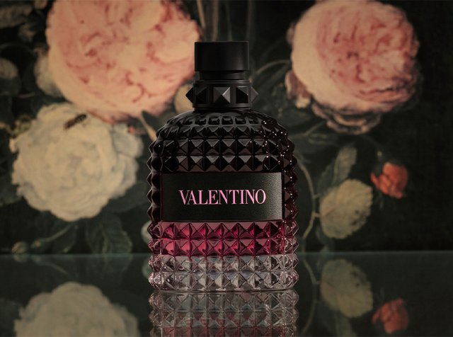 Bottiglia di profumo Valentino con design sfumato nero e rosa strutturato, posizionata su una superficie riflettente con sfondo floreale.