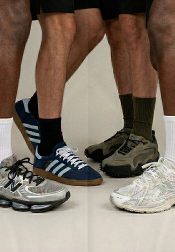 Quatre paires de jambes portant différentes baskets et chaussettes se tiennent sur un sol neutre devant un fond uni.