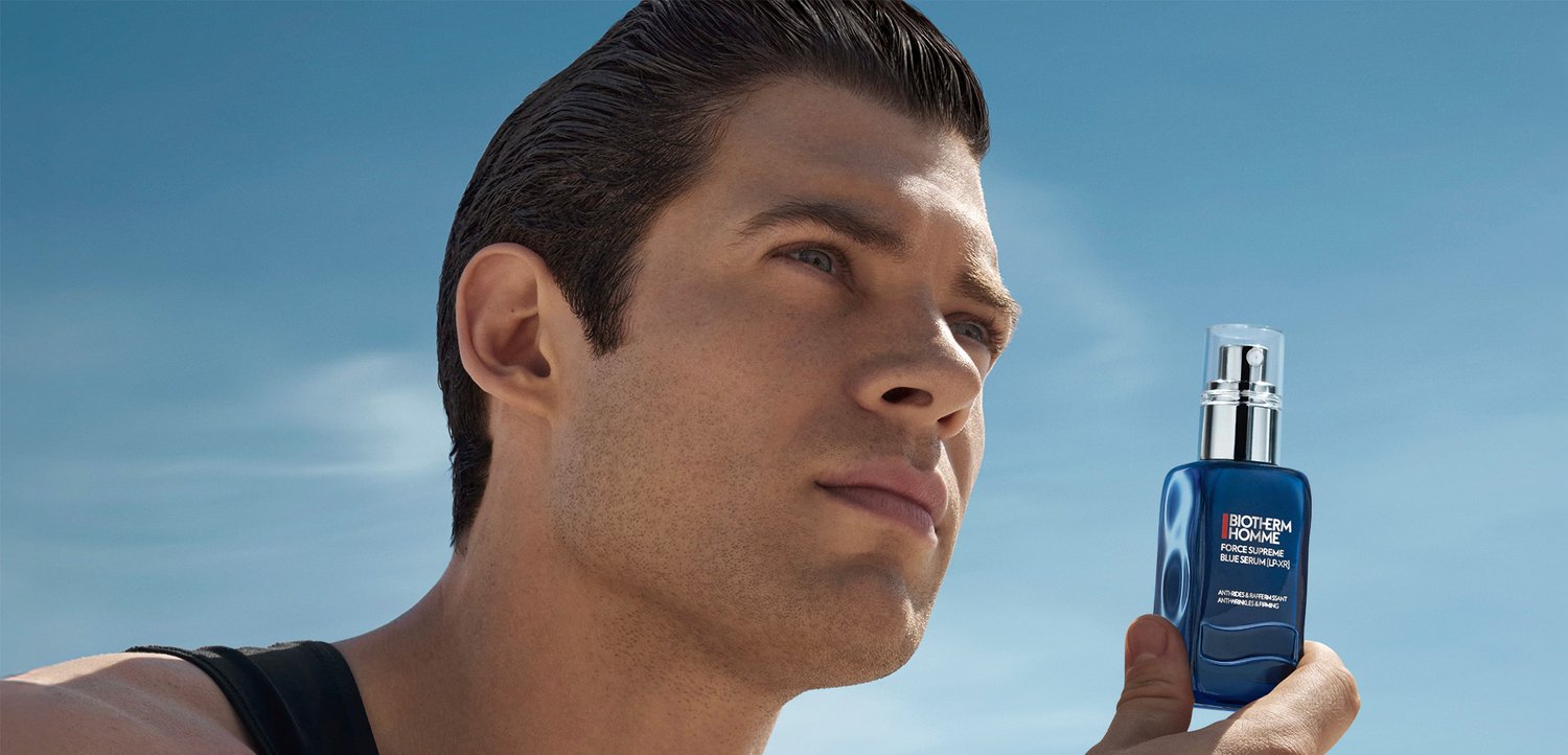 Ein Mann mit nach hinten gekämmtem Haar hält eine blaue Serumflasche vor einem klaren Himmel und vermittelt damit Selbstbewusstsein und Fokus auf Hautpflege.