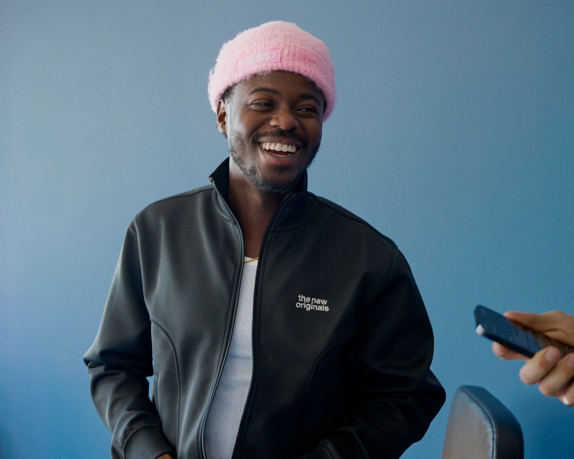 Veste noire zippée avec logo, débardeur blanc en dessous et bonnet en tricot rose pâle. Fond d'un mur bleu uni.