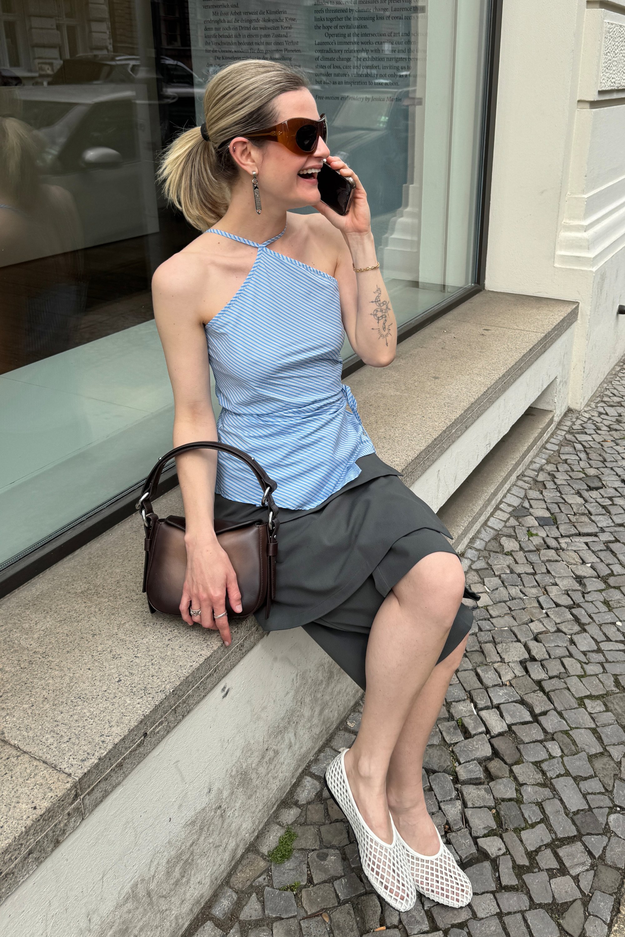 Donna con un ponytail, vestita con una maglietta a righe e una gonna grigia, seduta su un ledge di pietra, parla al telefono e tiene una borsa marrone.