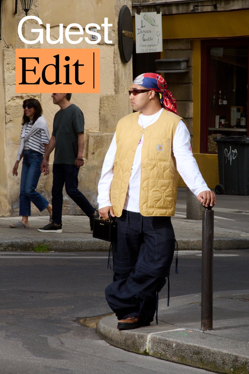 Un hombre con un chaleco amarillo y gafas de sol se apoya en un poste, sosteniendo una bolsa negra. Dos personas pasan caminando vestidas de manera informal. Entorno urbano.