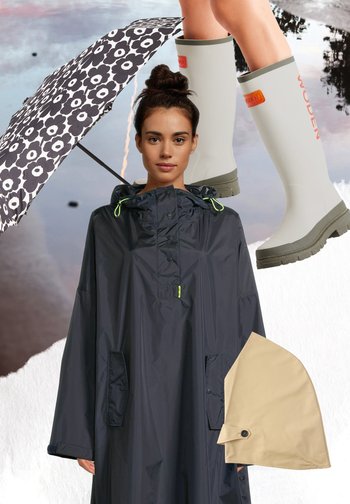 Frau trägt schwarzen Regenponcho, hält schwarz-weiß geblümten Regenschirm, beige abnehmbare Regenkapuze und weiße Gummistiefel mit grünen Sohlen.