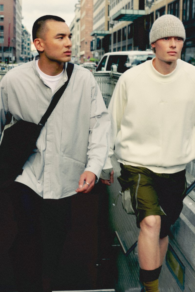 Twee jonge mannen lopen buiten. De ene draagt een gestreept overhemd met knopen en een zwarte crossbody tas; de andere draagt een witte sweatshirt en olijfgroene shorts.