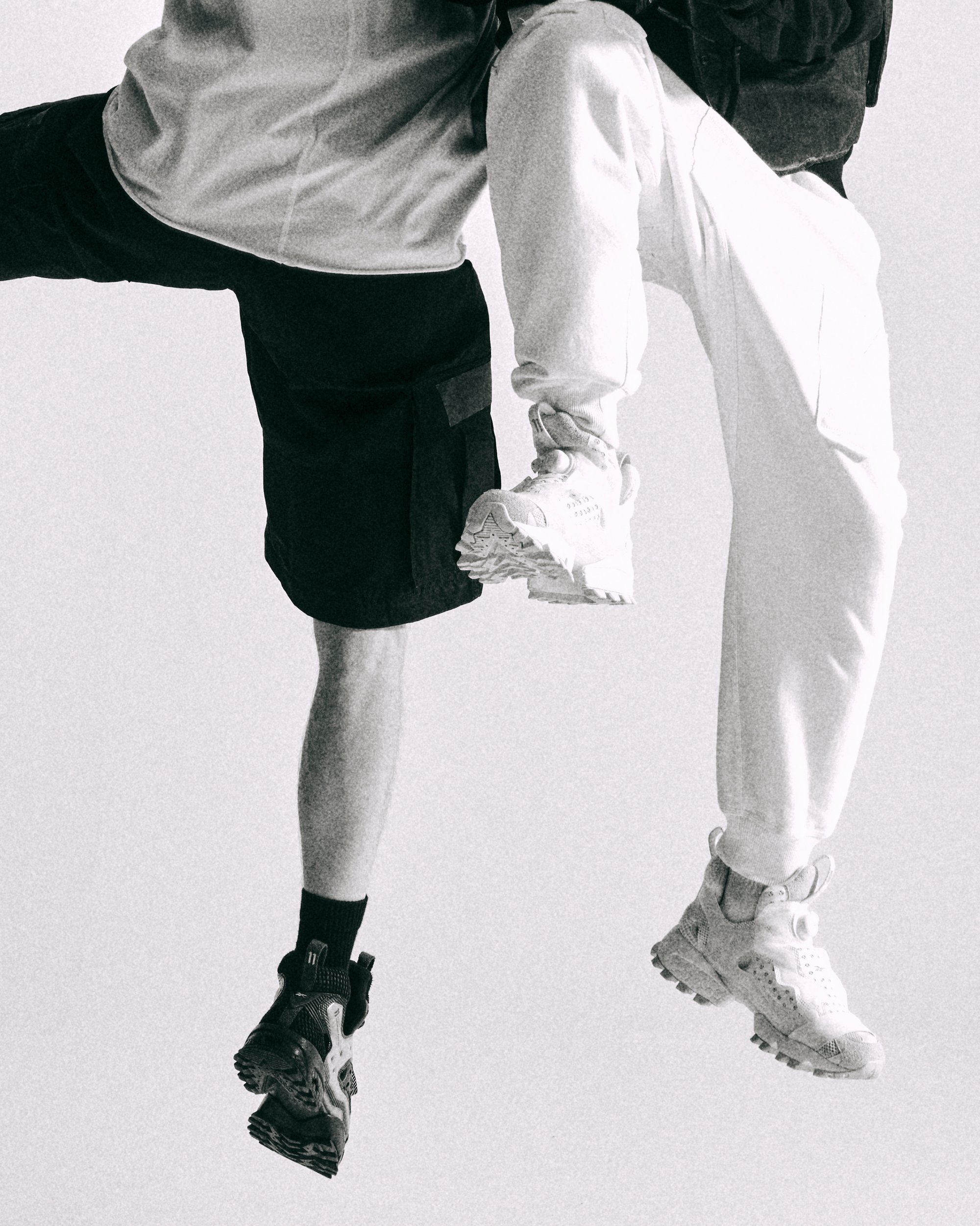 Two legs in motion, one wearing black shorts and black sneakers, the other in white pants and light sneakers, against a neutral background.