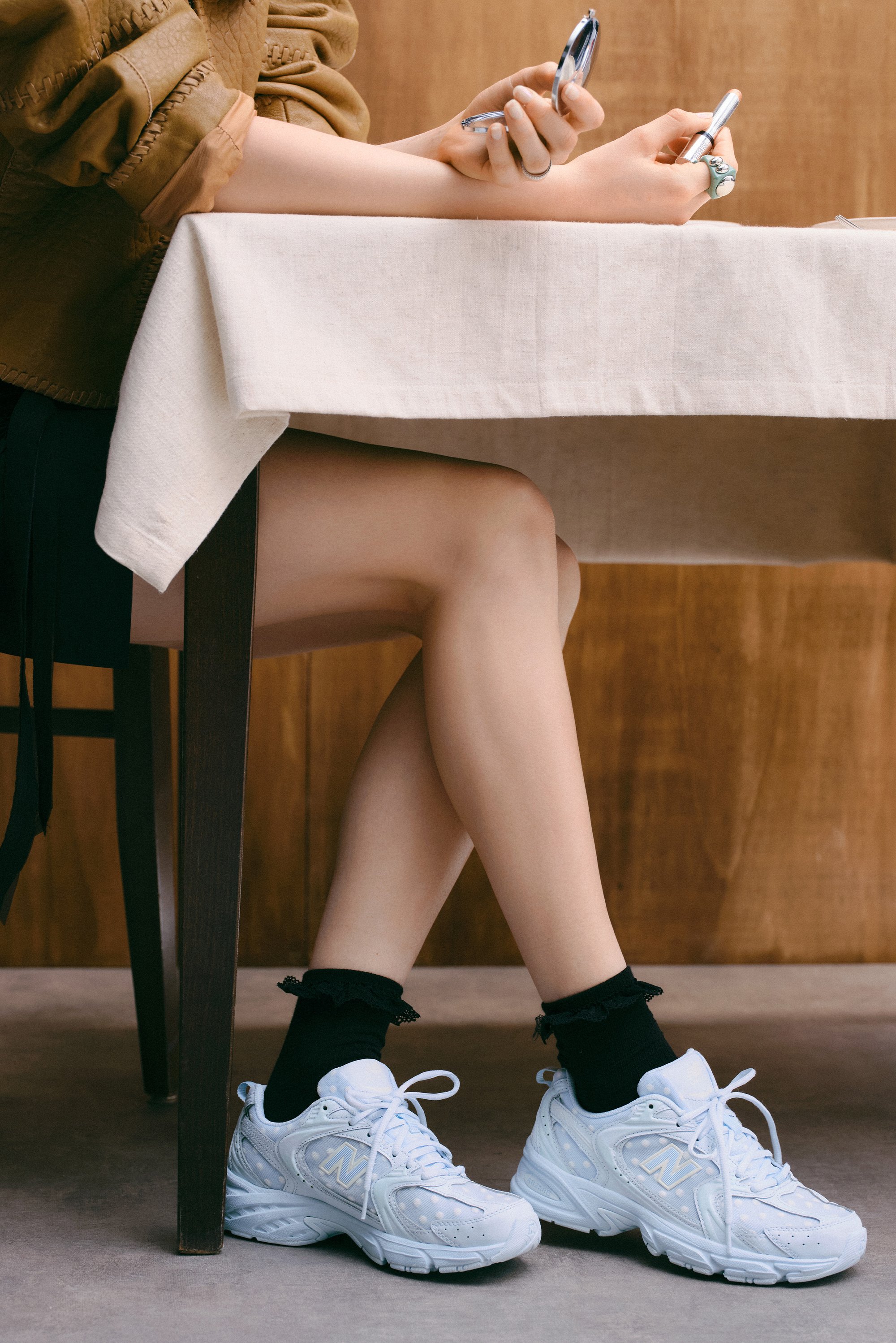Una persona è seduta a un tavolo, indossando una maglietta marrone e una gonna nera, con scarpe da ginnastica bianche e calze nere, mentre tiene in mano un telefono e uno specchio.