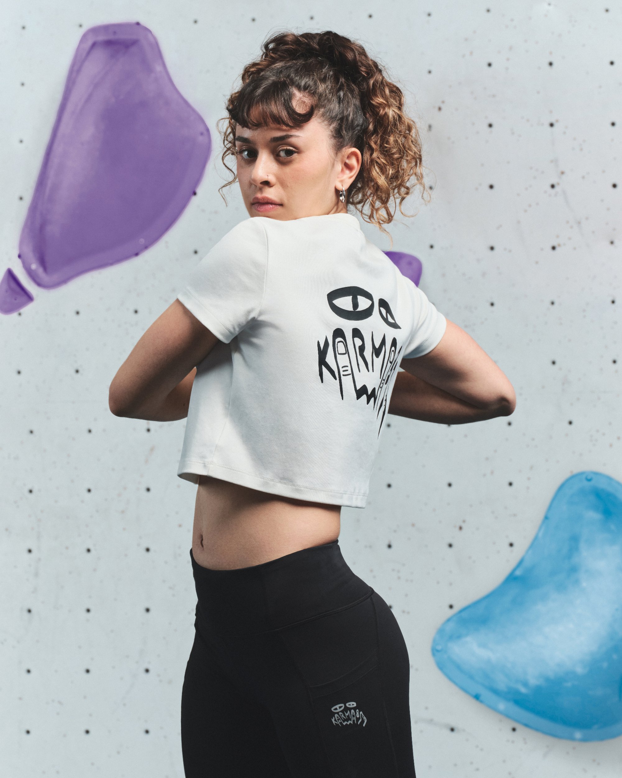 Camiseta corta blanca con un gráfico negro en la parte posterior, combinada con legging negros. El fondo presenta una pared de boulder con presas de colores.