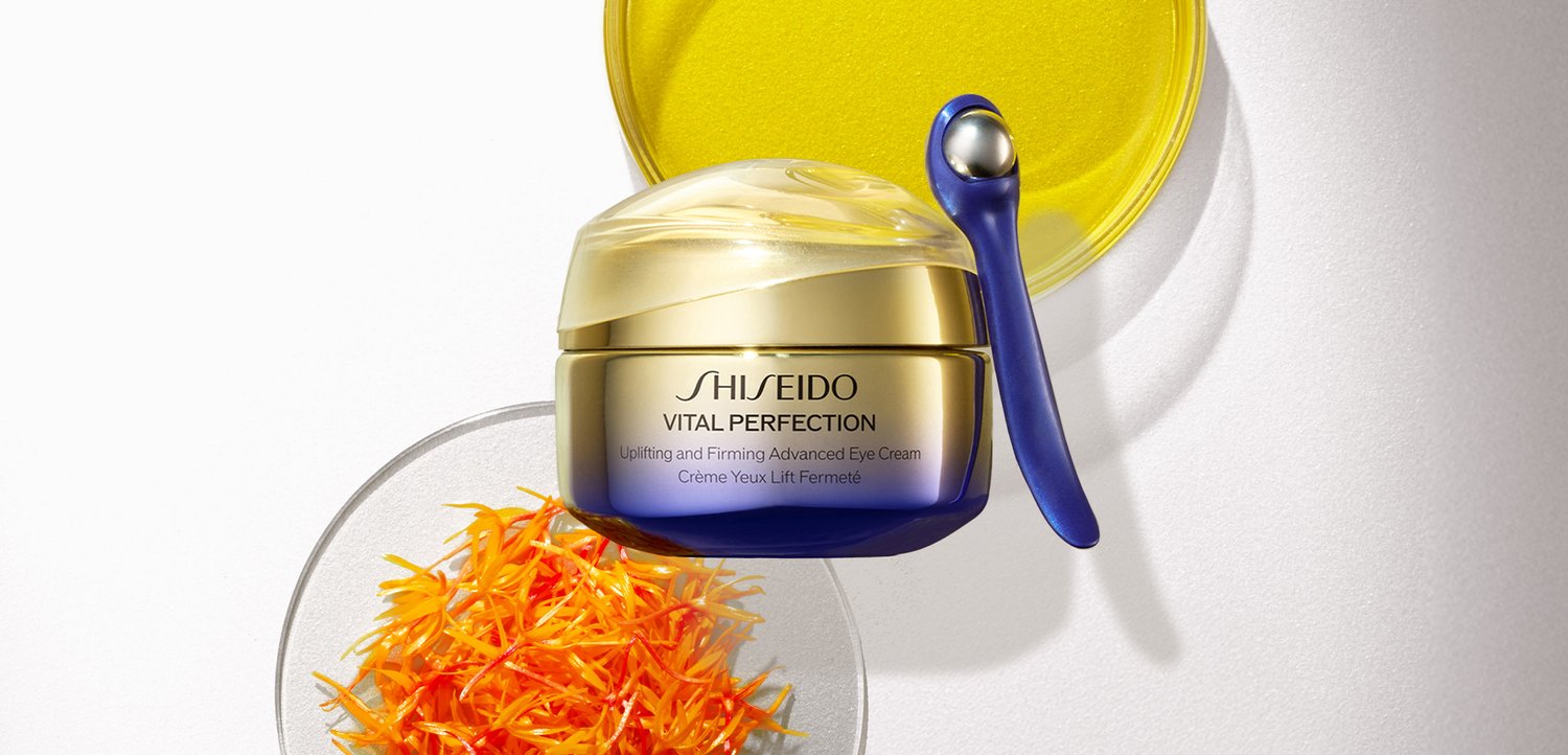 Shiseido Vital Perfection Augencreme-Tiegel mit einem kleinen blauen Applikator, zusammen mit gelben Blütenblättern auf runden Glasschalen vor weißem Hintergrund.