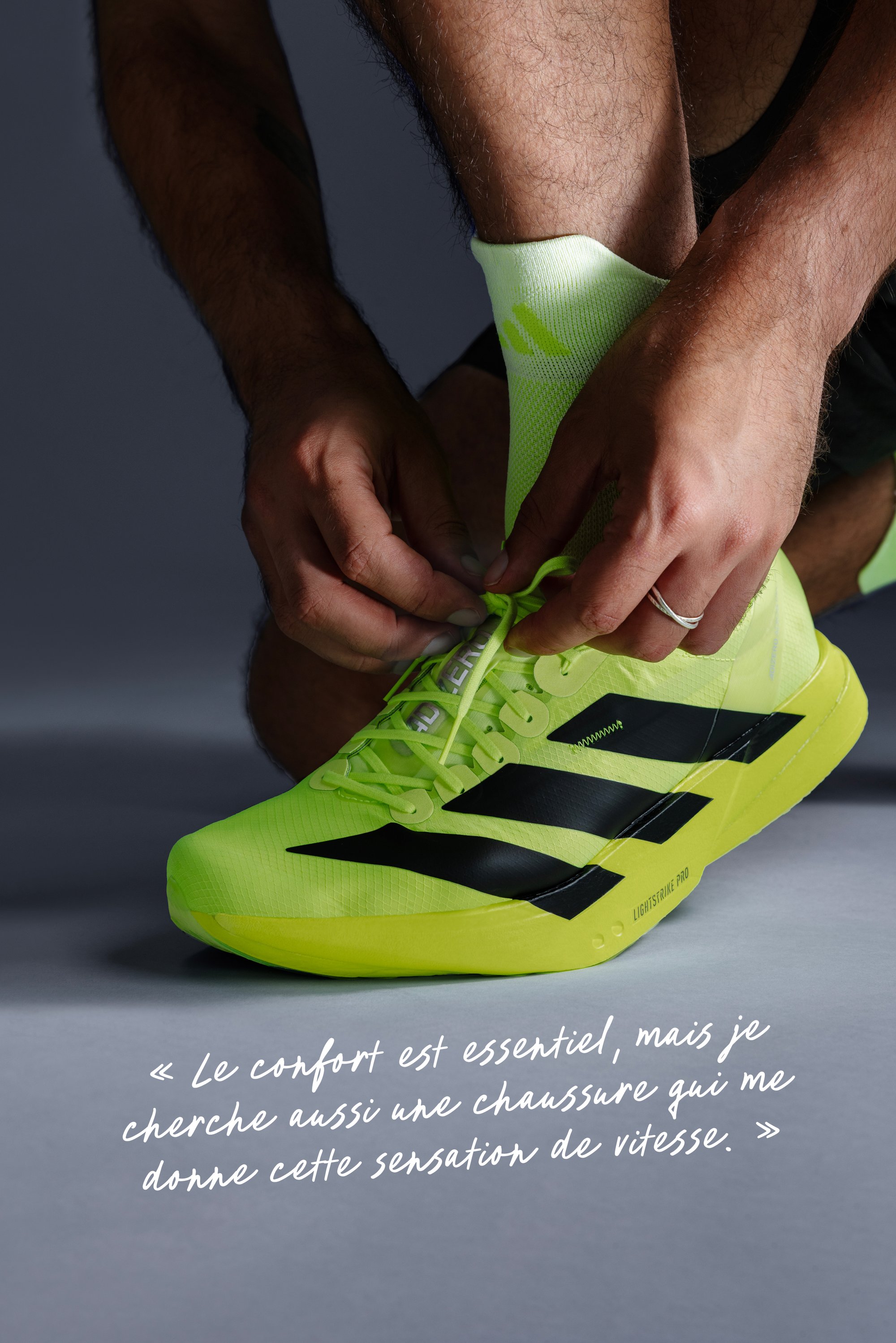 Mains nouant une chaussure de course Adidas jaune néon avec des rayures noires, gros plan sur le pied et les jambes inférieures sur fond uni.