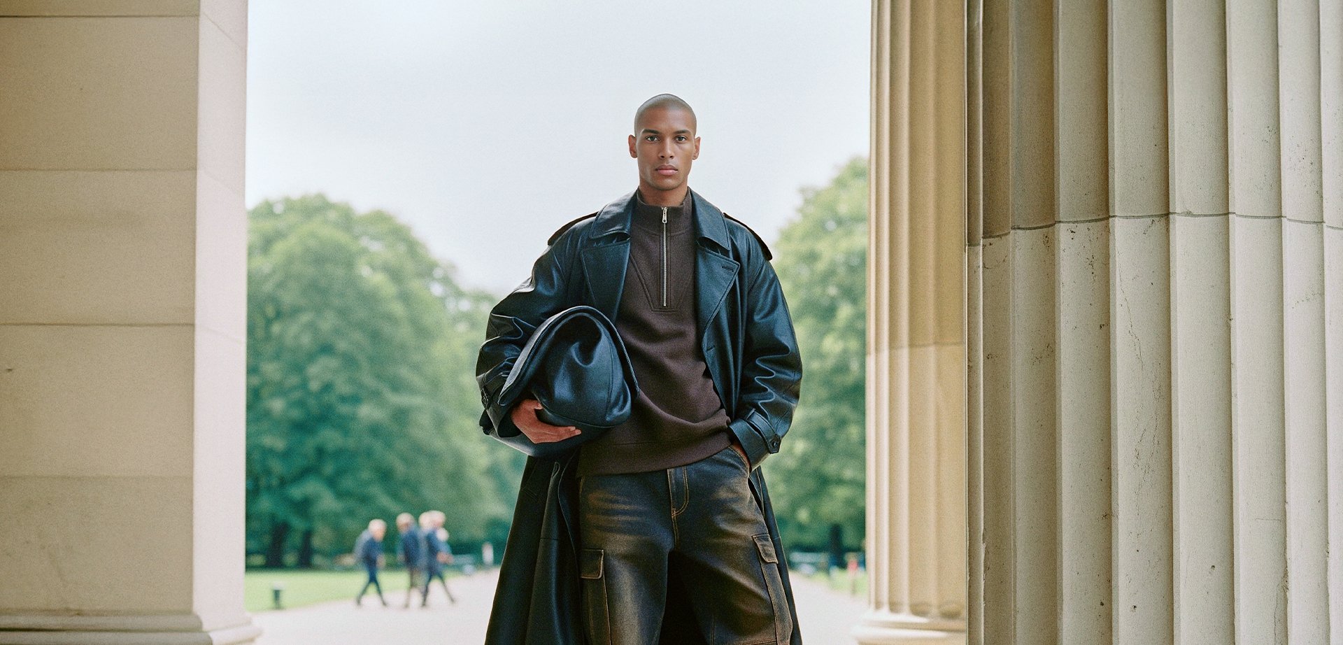 Black leather trench coat, dark brown high-collar top, black bag, and loose black trousers. Smooth textures, tailored fit, and subtle accents.