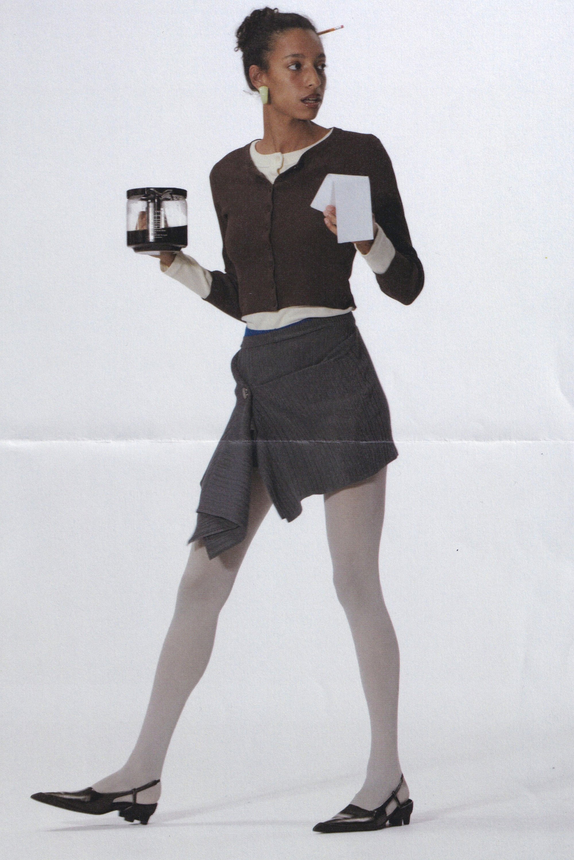 Bruine cardigan over een witte top, grijze wikkelrok en lichte maillot. Houdt een koffiepot en een witte kaart vast. Zwarte hakken, eenvoudige oorbellen.