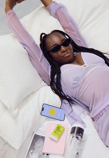 Een vrouw met gevlochten haar, die een zonnebril en een lavendelkleurige outfit draagt, ontspant op een bed omringd door een telefoon en schoonheidsproducten.