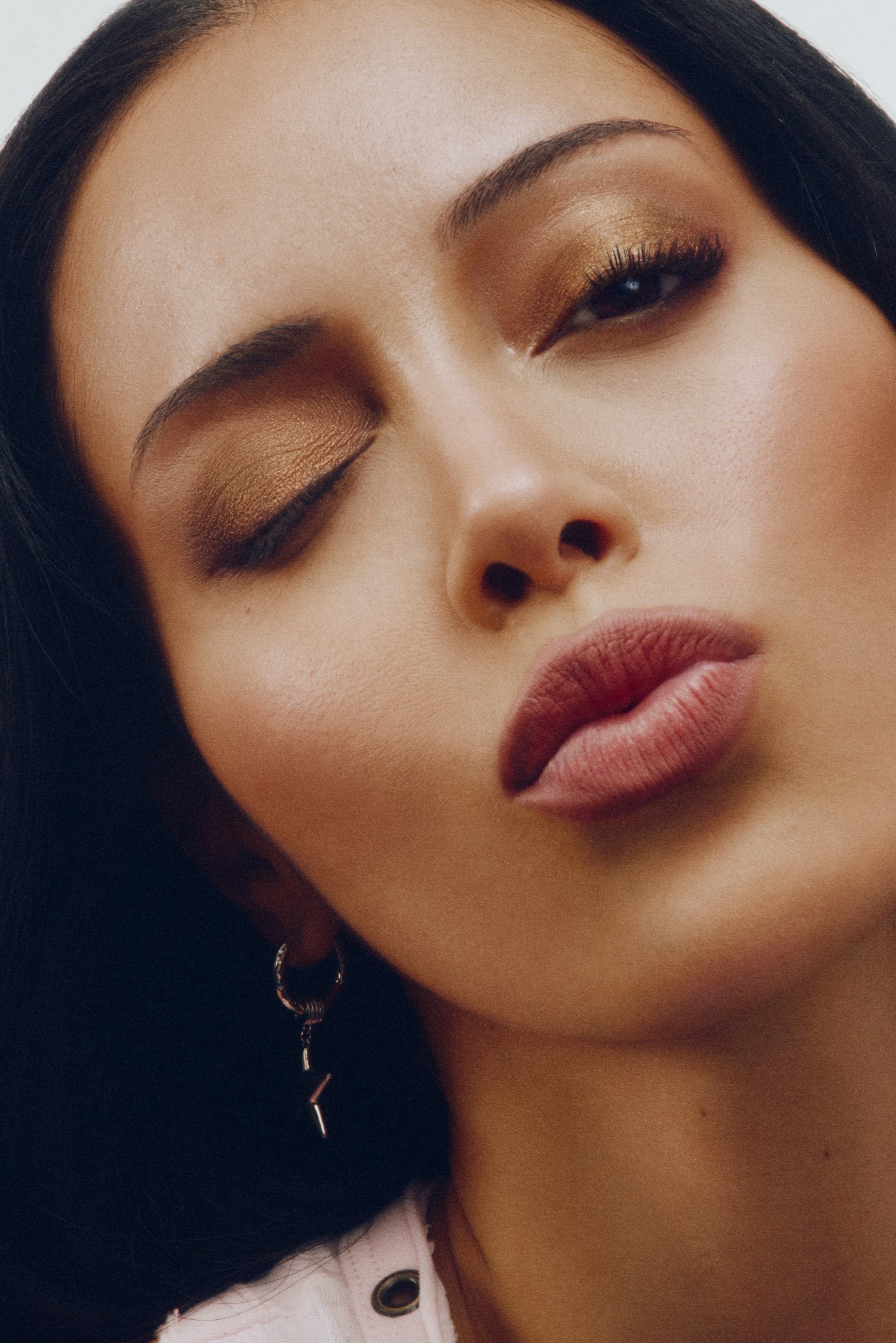 Primer plano de un rostro con sombra de ojos bronce, labios mate en un suave tono rosa y pendientes de aro plateados con un dije de rayo. Textura de piel suave.