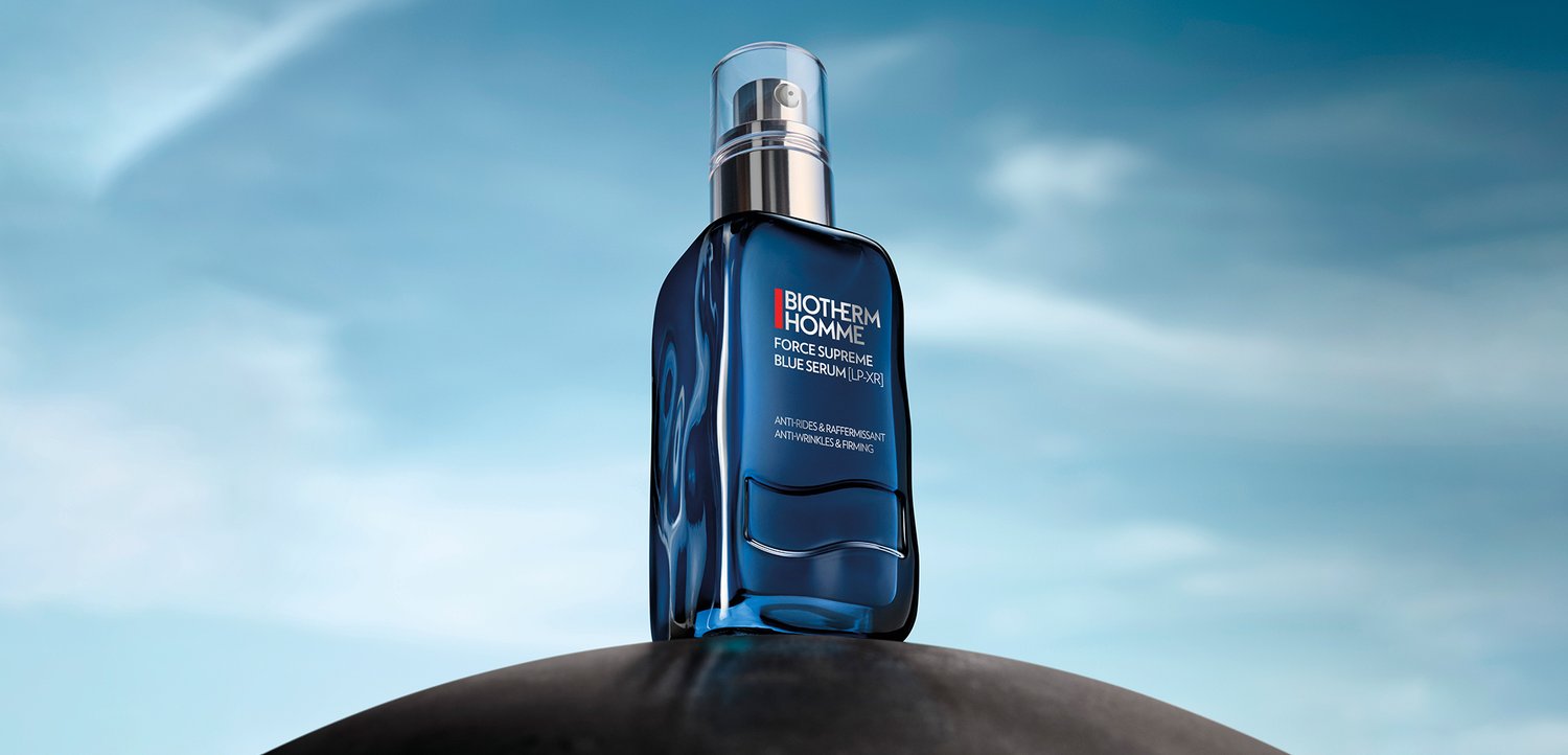 Blue serum bottle on a black surface, with a soft blue sky backdrop, highlighting the product's sleek design and branding.