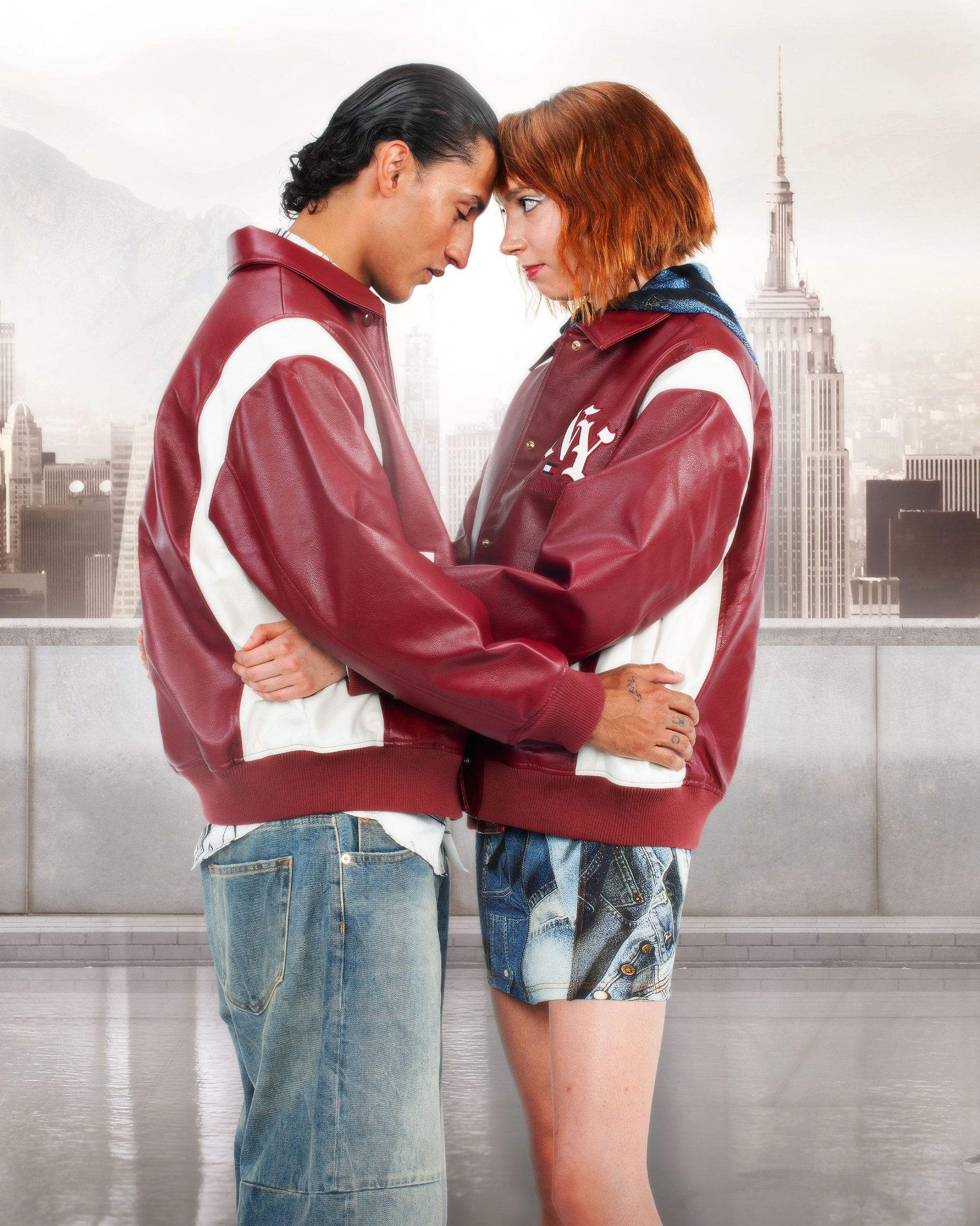 A man and woman in matching red and white jackets embrace, their foreheads touching, against a backdrop of a city skyline featuring the Empire State Building.