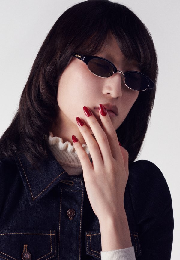 Occhiali da sole ovali neri con marchio dorato, indossati da una persona con capelli scuri. Unghie ovali rosse contrastano con una giacca di jeans e un colletto bianco a balze.