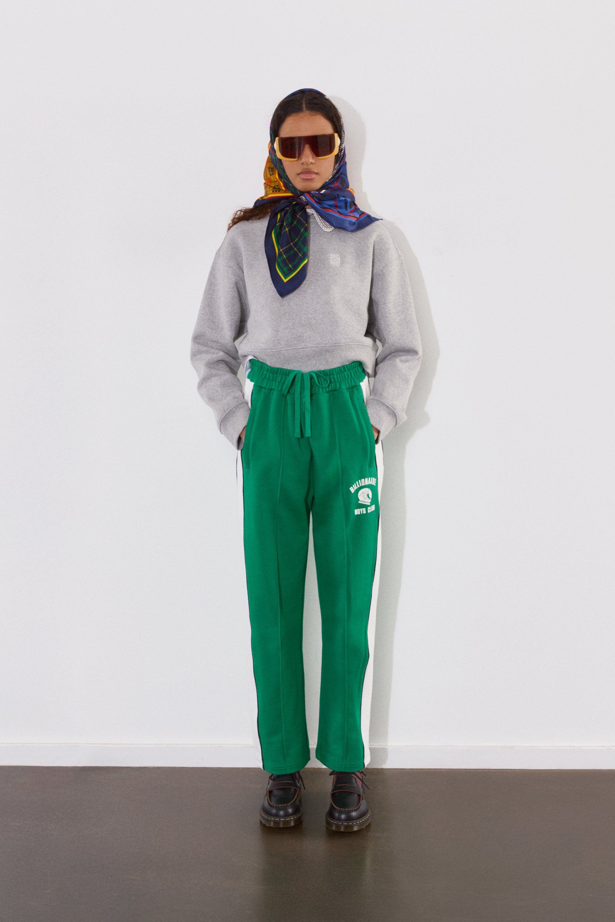 Una persona está de pie contra una pared blanca, vestida con un sudadera gris, pantalones deportivos verdes con un logotipo, gafas de sol y una bufanda con un patrón.