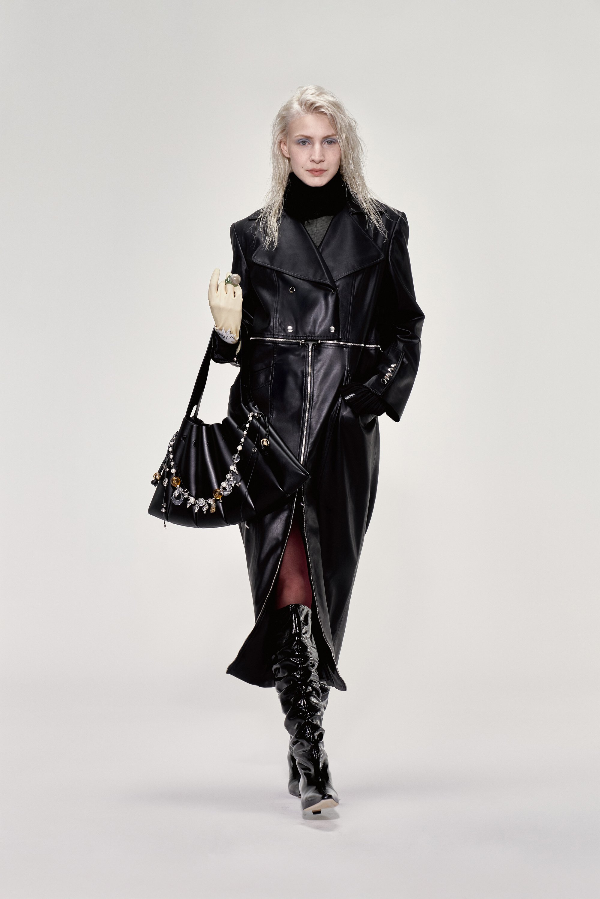 Modelo con un largo abrigo de cuero negro y botas altas negras hasta la rodilla sostiene un bolso elegante adornado con joyas. El fondo claro resalta la indumentaria.
