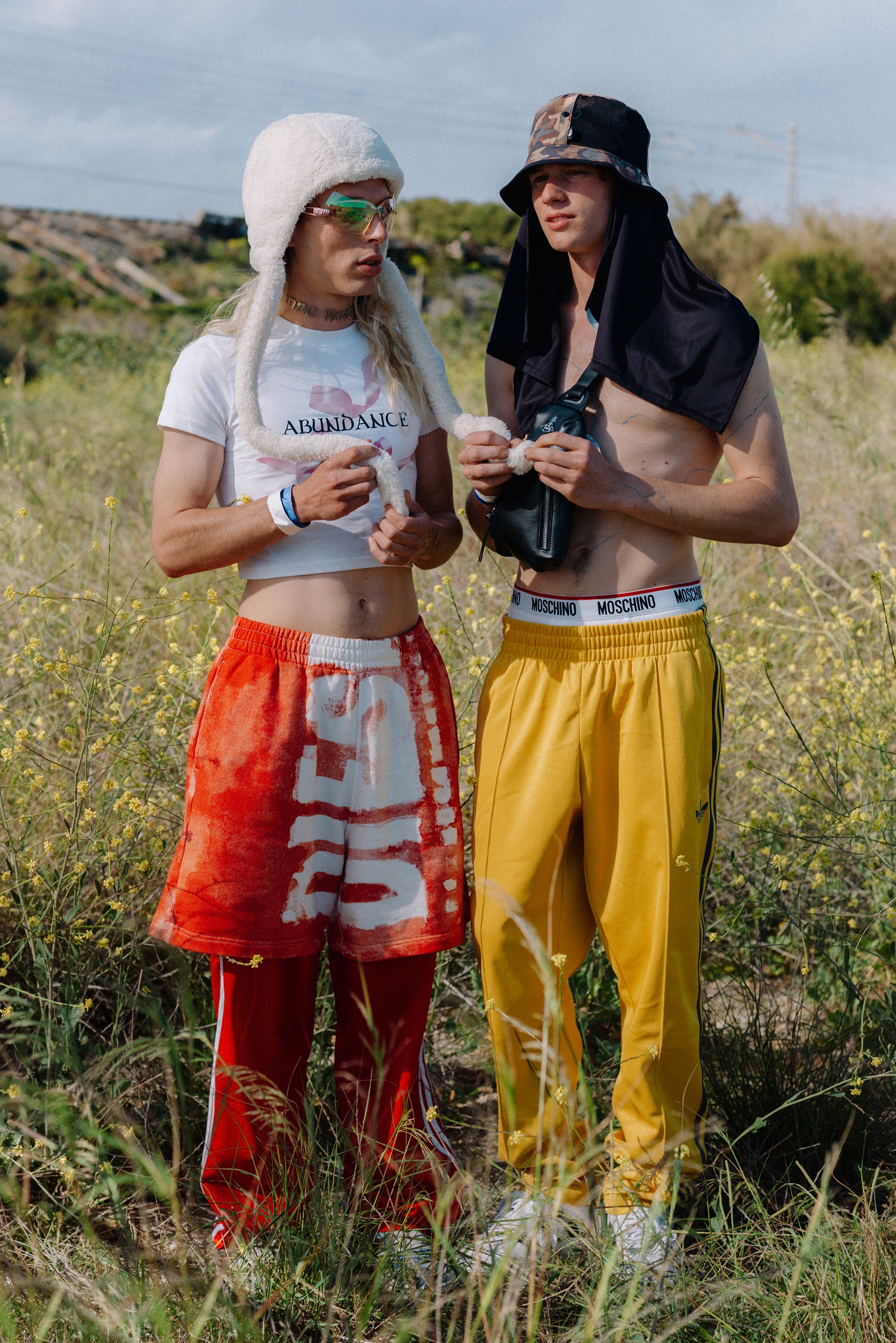 Twee jonge mannen staan in een grasveld. De één draagt een witte, harige hoed en een crop top, de ander is gekleed in een zwart shirt en gele broek; beide zijn zonder shirt.