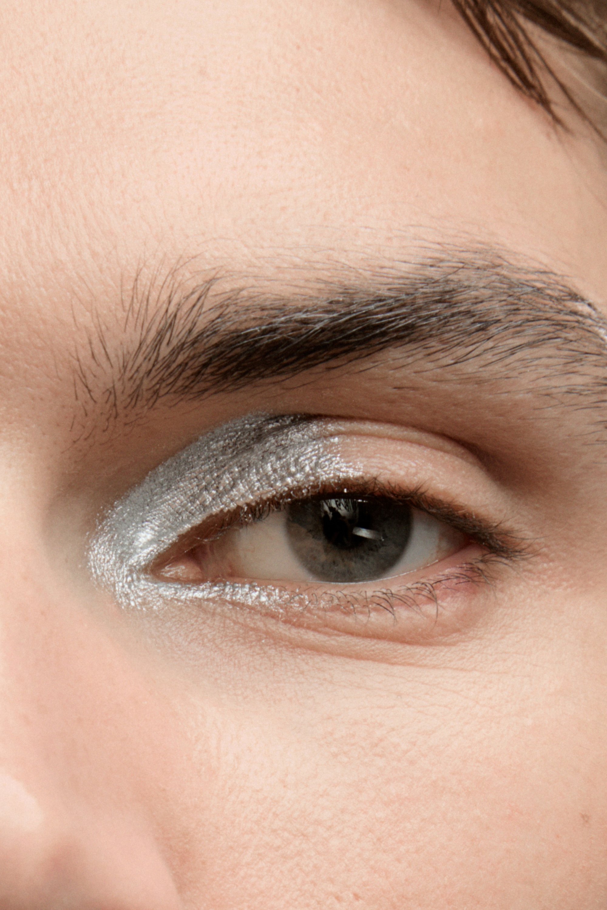 Primer plano de un ojo con sombra de ojos plateada que resalta el párpado, mostrando pestañas claras y una ceja definida. La piel parece suave y natural.