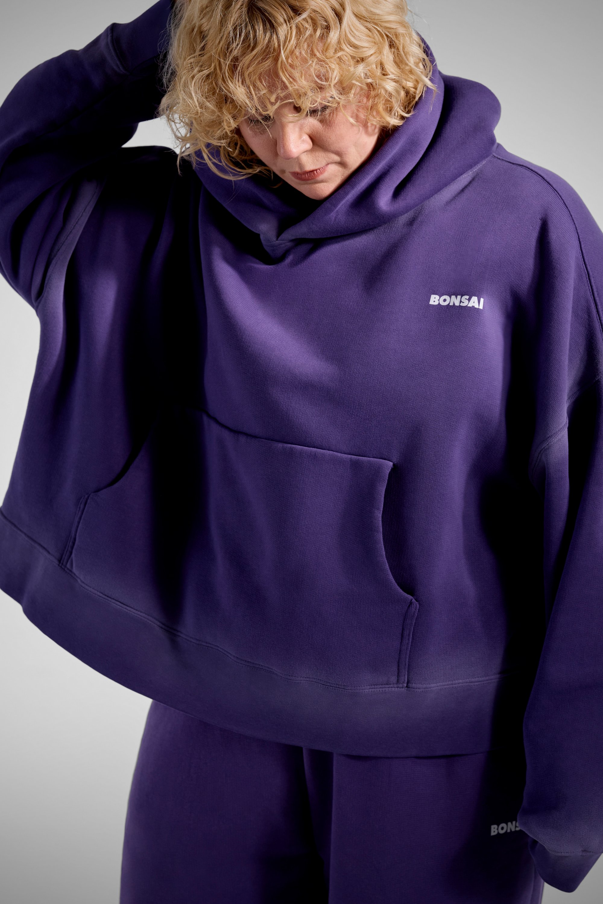 Henkilö, jolla on kiharat hiukset, käyttää hieman löysää violettia hupparia, jossa on etutasku. He näyttävät katsovan alas, mikä korostaa rentoa istuvuutta.